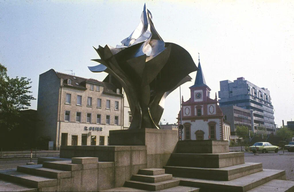 Flamme, Ansicht mit Häuserzeile und Kirchturm im Hintergrund, Skulptur, Edelstahl
